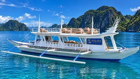 A white-and-blue Velaya outrigger boat with an upper deck on turquoise water beside limestone islands.