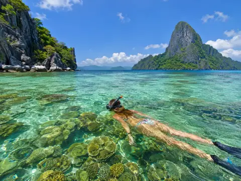 A snorkeler floating above a shallow coral reef with limestone islands in the background.
