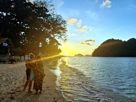 Travelers standing at the water's edge as the sun sets behind the islands on a Palawan beach.